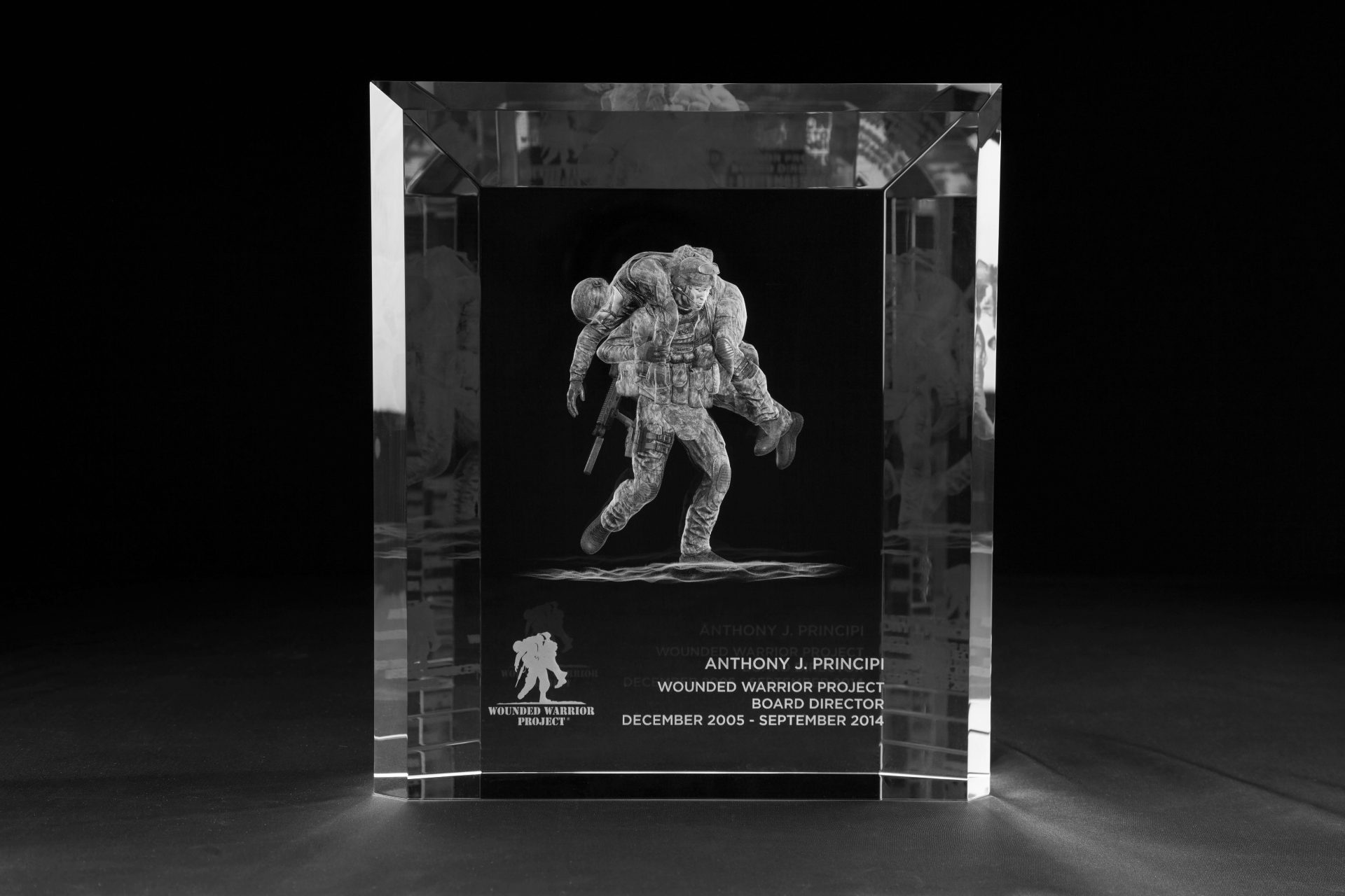 The piece is a large-format optical crystal block, bevelled at every edge, with the Wounded Warrior Project's defining image: a soldier carrying his fallen brother, engraved inside the crystal 3-dimentionally with the kind of depth and detail that stops a conversation in a room. Below the figure sits the organization's logo and a single inscription: Board Director, December 2005 – September 2014 — six words that name the role, mark the nine years, and say to one specific person. This is the project we designed for veterans service organization.