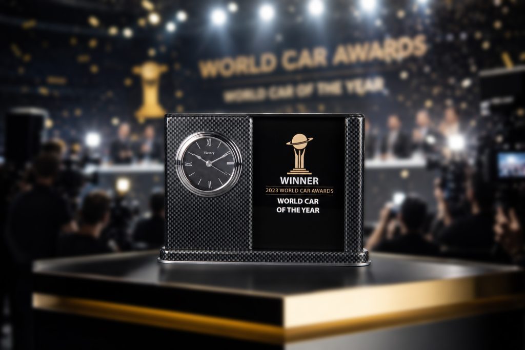 A premium World Car Awards trophy is displayed prominently on a sleek black podium with gold trim during a championship press conference. The award features a carbon-fiber-patterned frame, an integrated silver clock face, and a glossy black center panel engraved with the World Car Awards logo and “Winner – World Car of the Year.” In the softly blurred background, photographers, television cameras, bright stage lights, and falling gold confetti create a celebratory atmosphere. Dramatic spotlights and shallow depth of field draw attention to the award, emphasizing its prestige and significance in a luxury, high-end editorial photography style. Crystal award made by Crystal Sensations