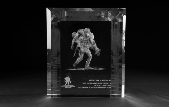 The piece is a large-format optical crystal block, bevelled at every edge, with the Wounded Warrior Project's defining image: a soldier carrying his fallen brother, engraved inside the crystal 3-dimentionally with the kind of depth and detail that stops a conversation in a room. Below the figure sits the organization's logo and a single inscription: Board Director, December 2005 – September 2014 — six words that name the role, mark the nine years, and say to one specific person. This is the project we designed for veterans service organization.