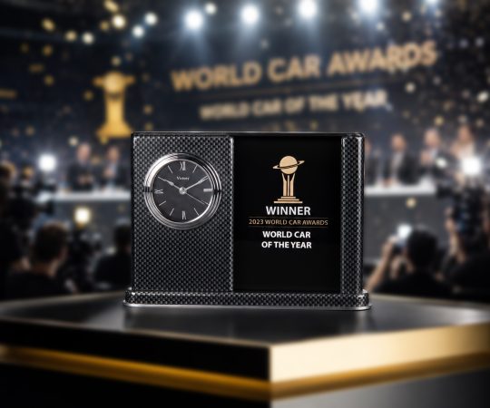 A premium World Car Awards trophy is displayed prominently on a sleek black podium with gold trim during a championship press conference. The award features a carbon-fiber-patterned frame, an integrated silver clock face, and a glossy black center panel engraved with the World Car Awards logo and “Winner – World Car of the Year.” In the softly blurred background, photographers, television cameras, bright stage lights, and falling gold confetti create a celebratory atmosphere. Dramatic spotlights and shallow depth of field draw attention to the award, emphasizing its prestige and significance in a luxury, high-end editorial photography style. Crystal award made by Crystal Sensations